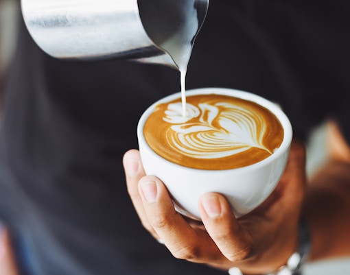 Barista pouring milk into a latte with art design in a white cup.