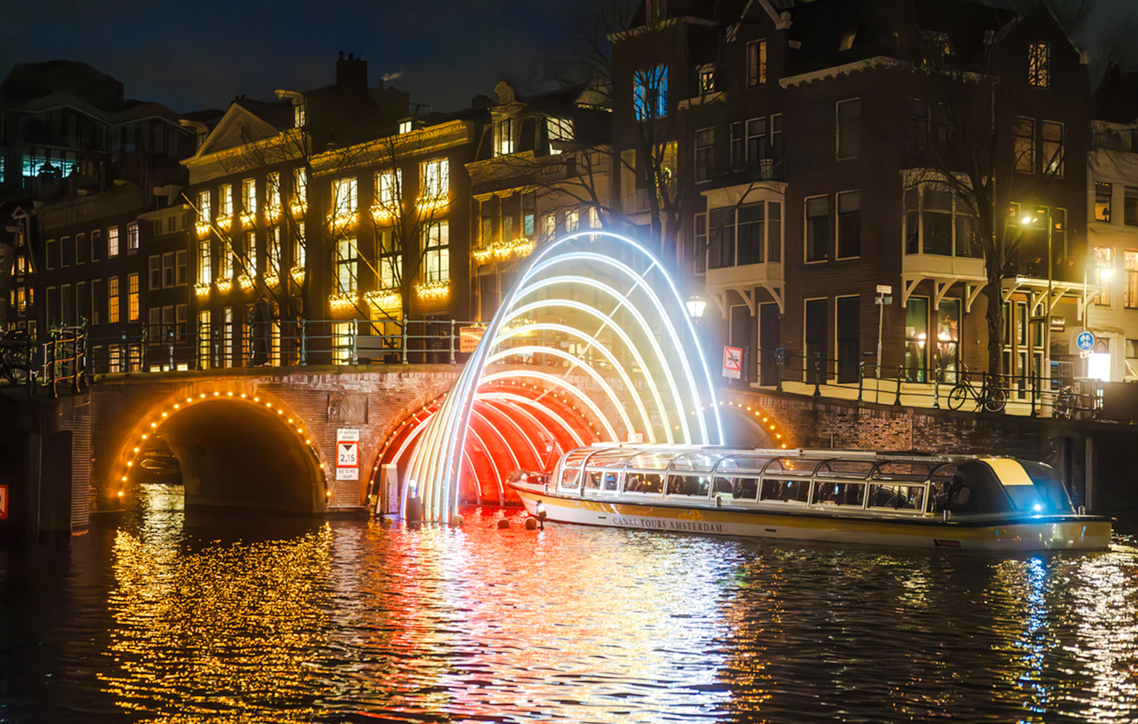 Canal cruise passing under illuminated arches during Amsterdam Light Festival.