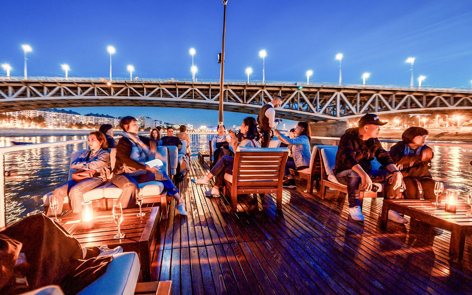 Guests on Budapest Danube River nighttime cruise with city lights reflecting on water.