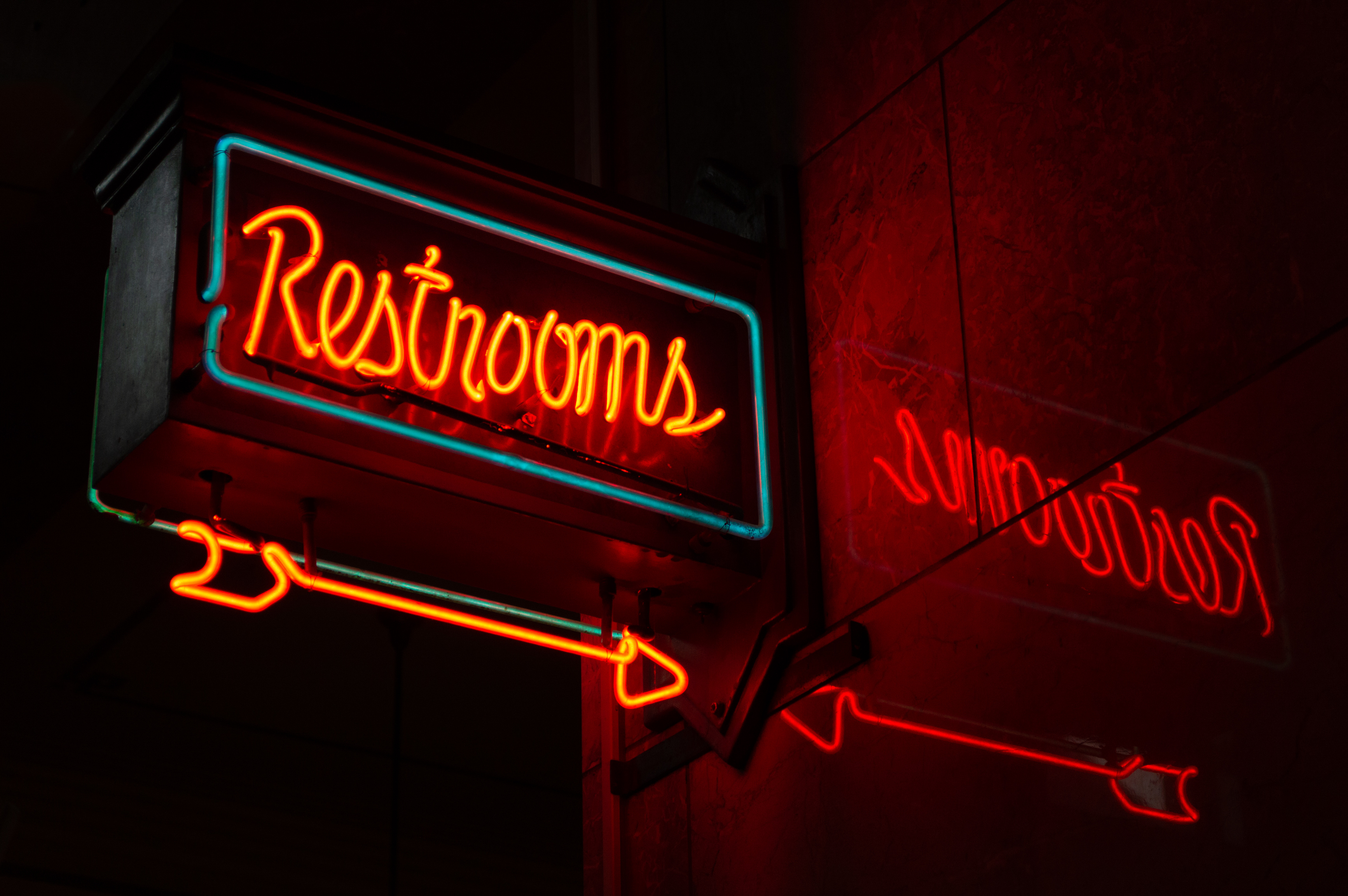 Neon restrooms sign with reflection in a dimly lit urban setting.