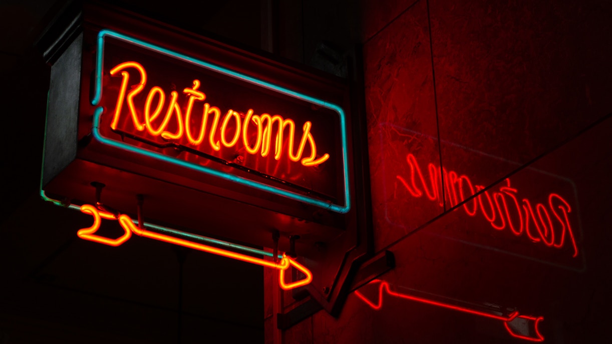 Neon restrooms sign with reflection in a dimly lit urban setting.