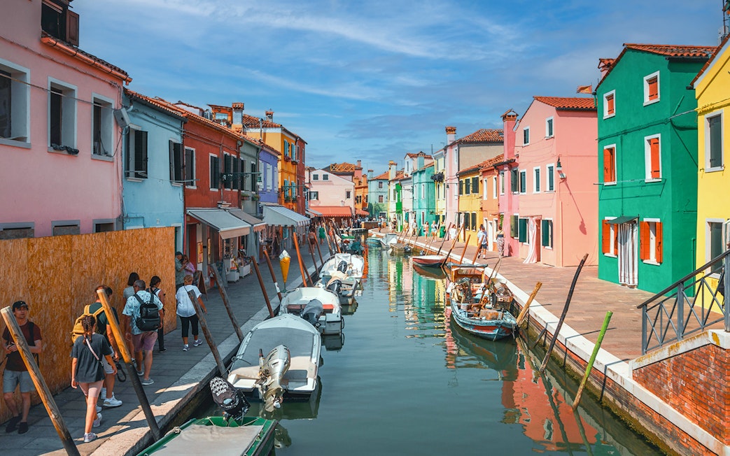 Colorful houses along Burano canal with people walking and boats docked.