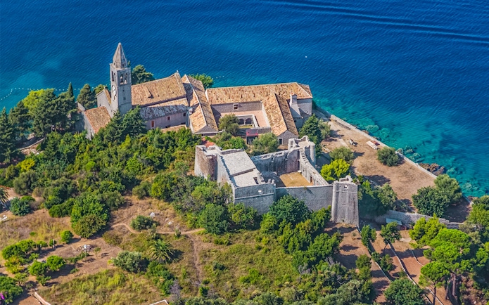 Aerial view of a historic monastery on the Elaphiti Islands near Dubrovnik, surrounded by lush greenery and blue sea.