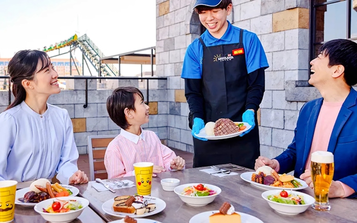 Server bringing food to a family at LEGOLAND Japan Nagoya with a roller coaster in the background.