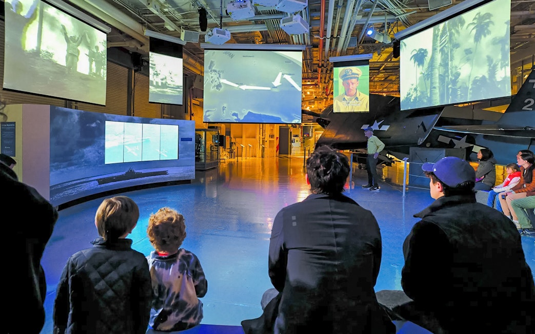 Visitors watching historical footage at a museum exhibit with multiple screens.