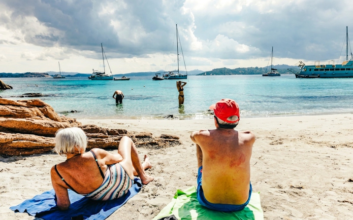Beachgoers relaxing on the shore with sailboats in La Maddalena Archipelago.