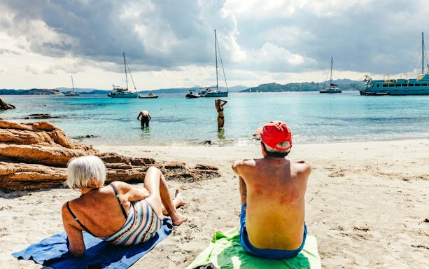 Beachgoers relaxing on the shore with sailboats in La Maddalena Archipelago.