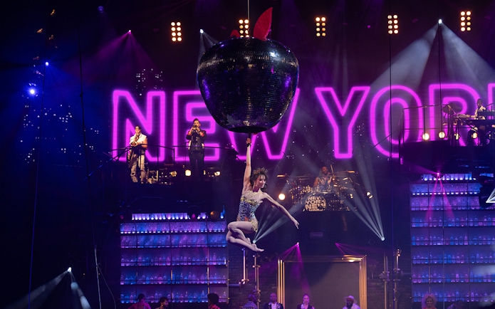 Performer hanging from a giant apple with "New York" sign in the background at Mad Apple show.