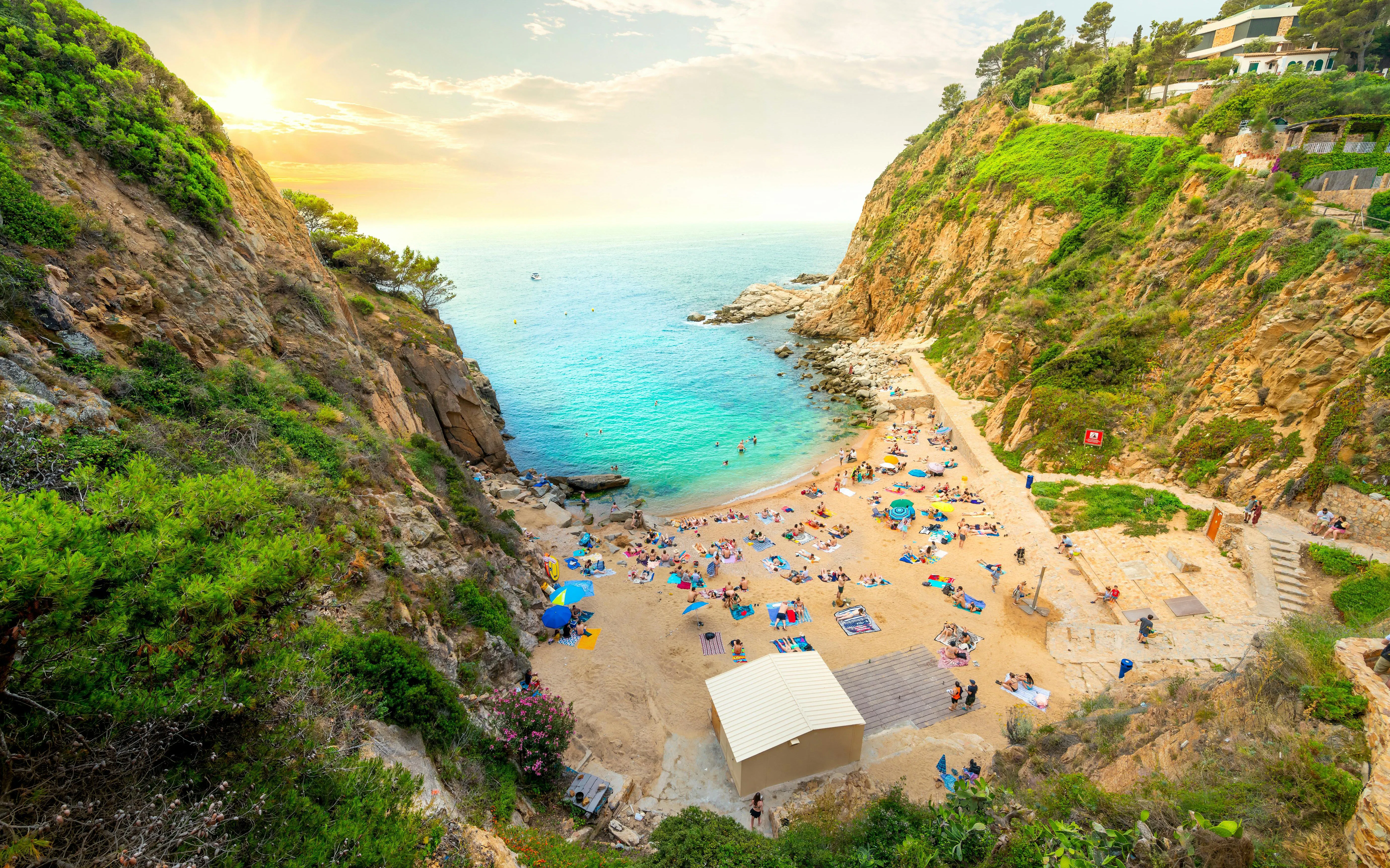 Tossa de Mar beach cove with sunbathers and turquoise water surrounded by cliffs.