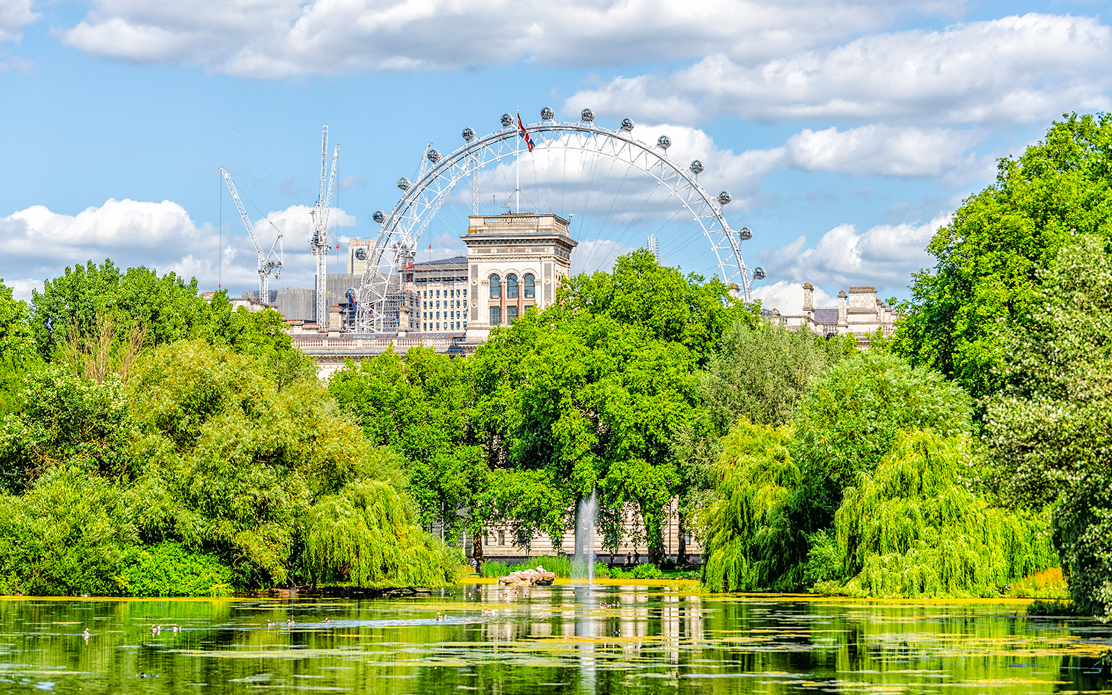 London Eye behind St James's Park lake and fountain, surrounded by greenery on a summer day.