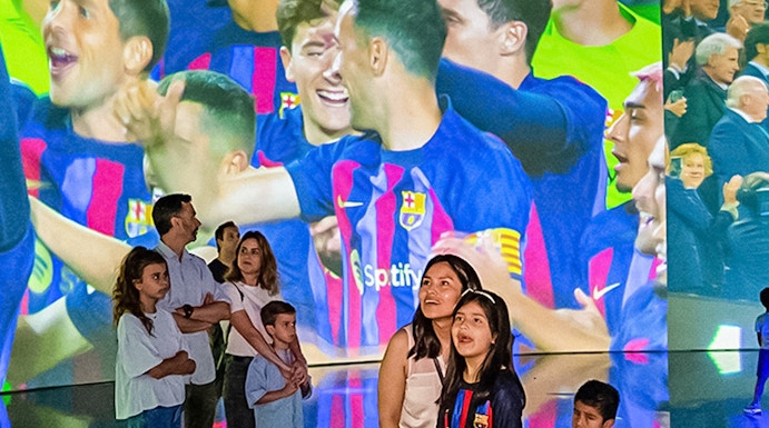 Visitors at Camp Nou viewing a large screen displaying FC Barcelona players celebrating.