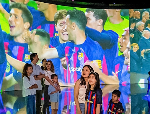 Visitors at Camp Nou viewing a large screen displaying FC Barcelona players celebrating.