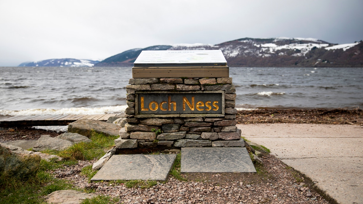 Loch Ness sign with view of the lake and snow-capped hills from Dores Beach.