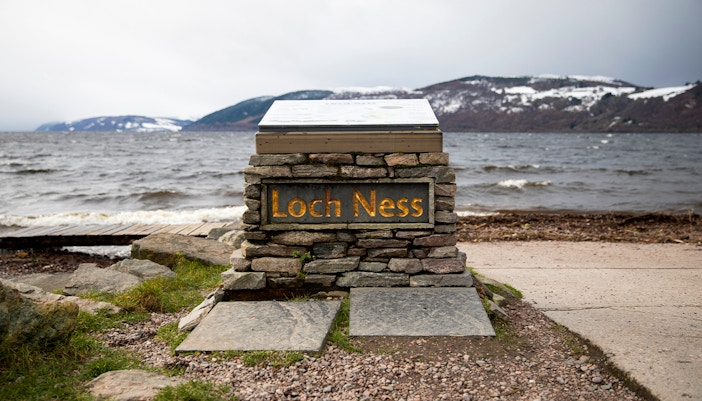 Loch Ness sign with view of the lake and snow-capped hills from Dores Beach.