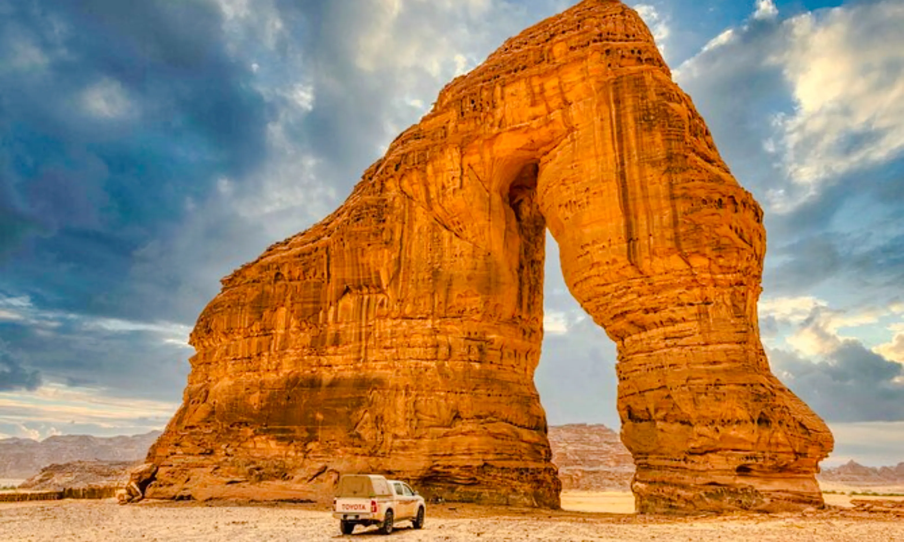 Elephant Rock in AlUla, Saudi Arabia, with a vehicle parked nearby under a cloudy sky.