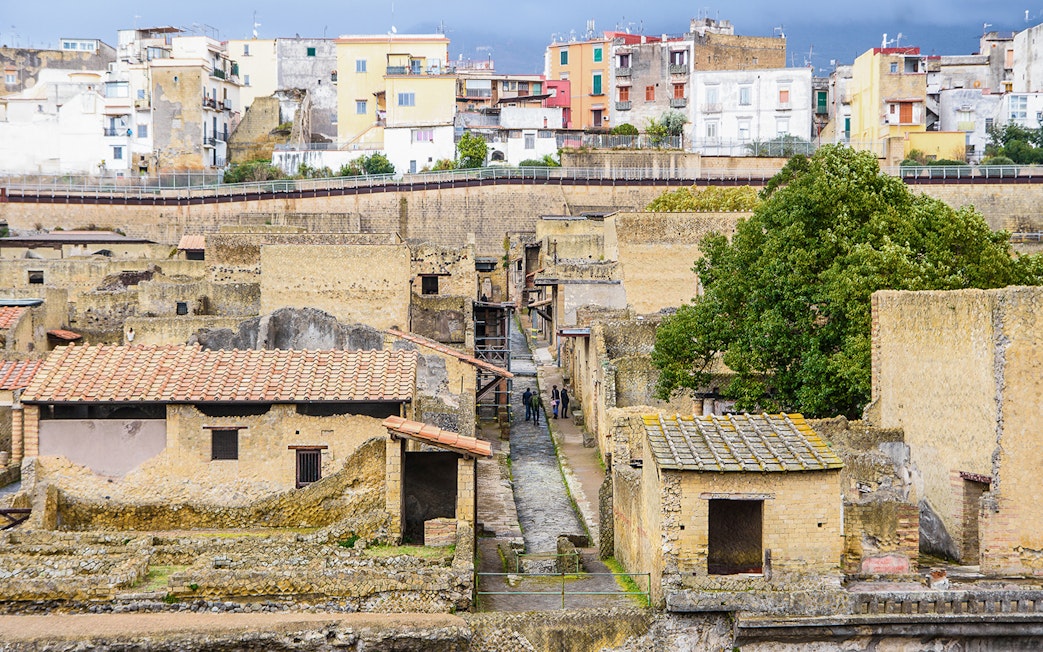 Visitors exploring ancient ruins at Herculaneum, Italy, with colorful buildings in the background.