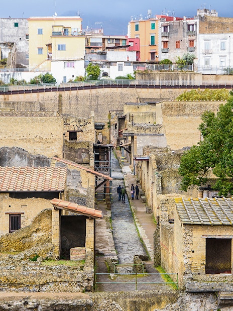 Visitors exploring ancient ruins at Herculaneum, Italy, with colorful buildings in the background.