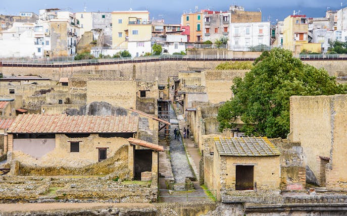 Visitors exploring ancient ruins at Herculaneum, Italy, with colorful buildings in the background.