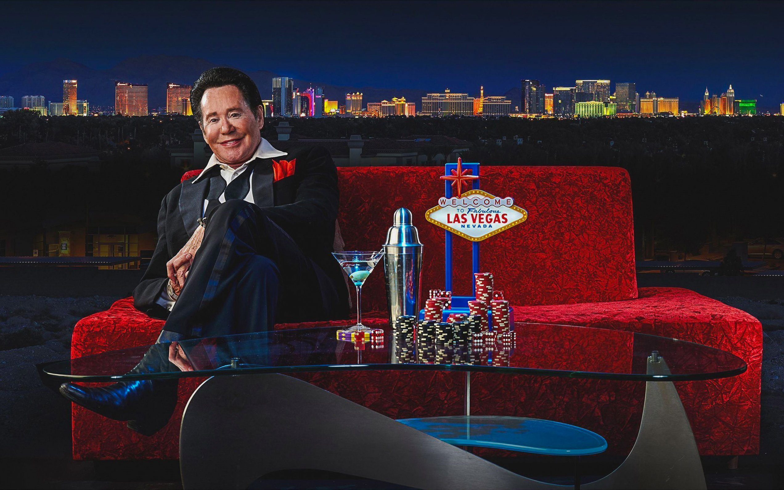 Wayne Newton on red couch with Las Vegas skyline, poker chips, and cocktail.