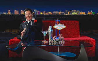 Wayne Newton on red couch with Las Vegas skyline, poker chips, and cocktail.