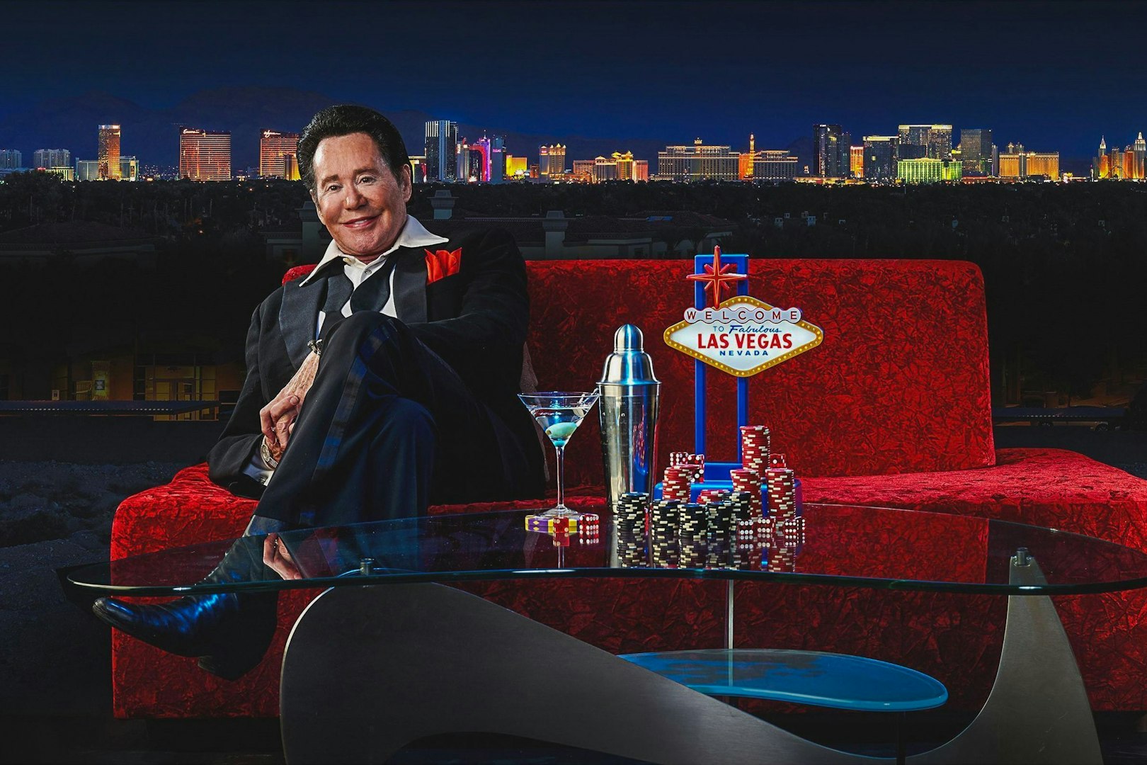 Wayne Newton on red couch with Las Vegas skyline, poker chips, and cocktail.