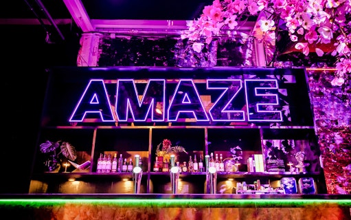 Neon sign at AMAZE Amsterdam with bar and floral decor.