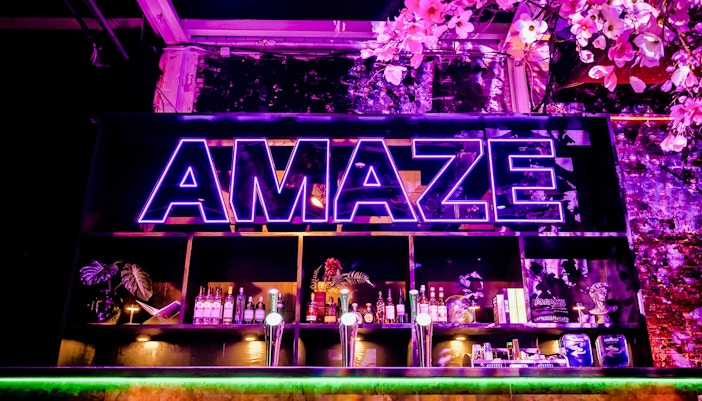 Neon sign at AMAZE Amsterdam with bar and floral decor.