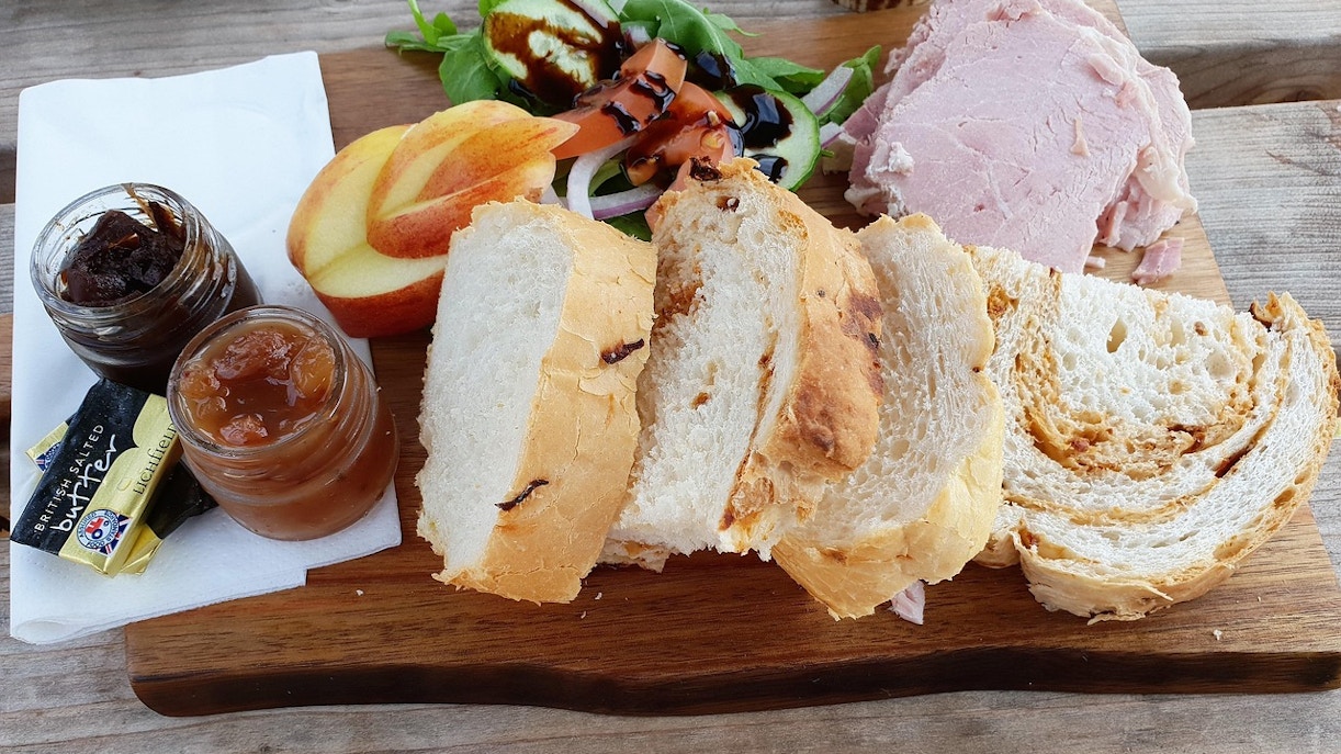 Ploughman's lunch with sliced bread, ham, apple, salad, and chutneys on a wooden board.