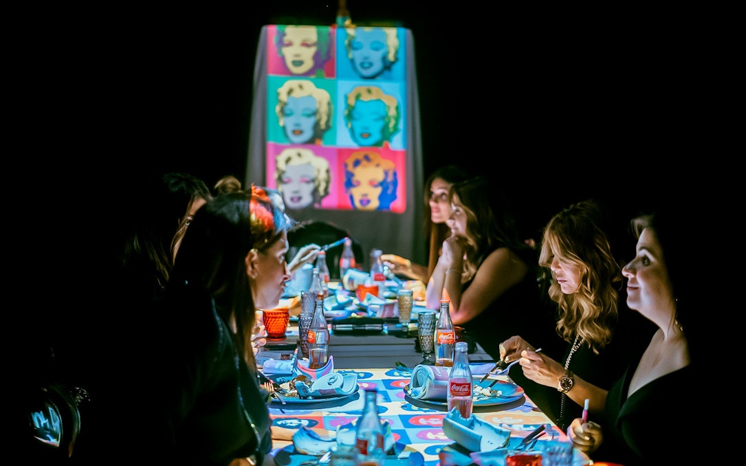 Guests dining at a table with pop art projections during the Seven Paintings Immersive Dining Show.