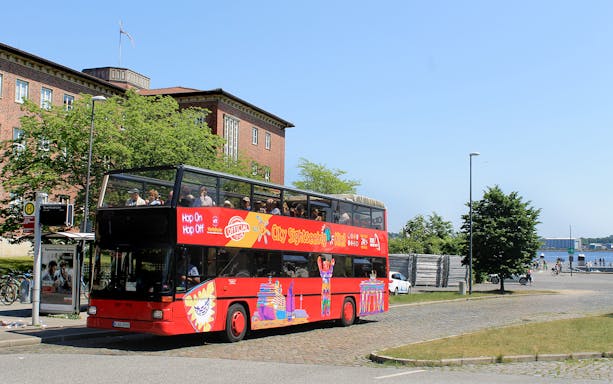 Red double-decker bus on Kiel Hop-On Hop-Off Tour near waterfront and historic building.