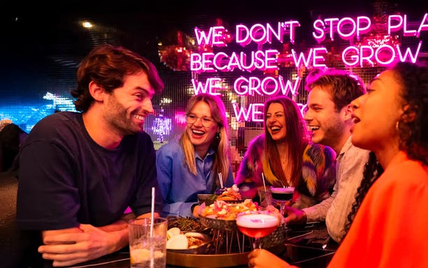 Guests enjoying food and drinks at All Out Amsterdam with neon sign in background.