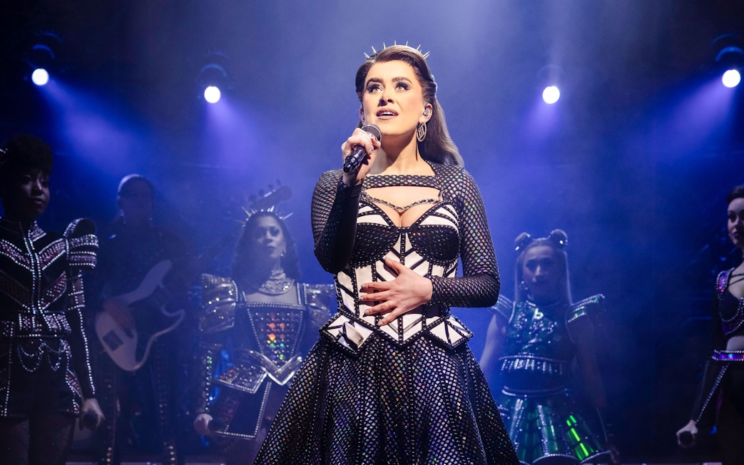 Performer singing on stage during the musical "Six" with a spotlight and backup singers.