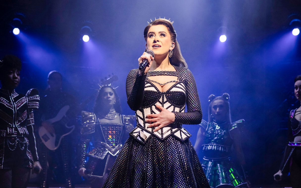 Performer singing on stage during the musical "Six" with a spotlight and backup singers.