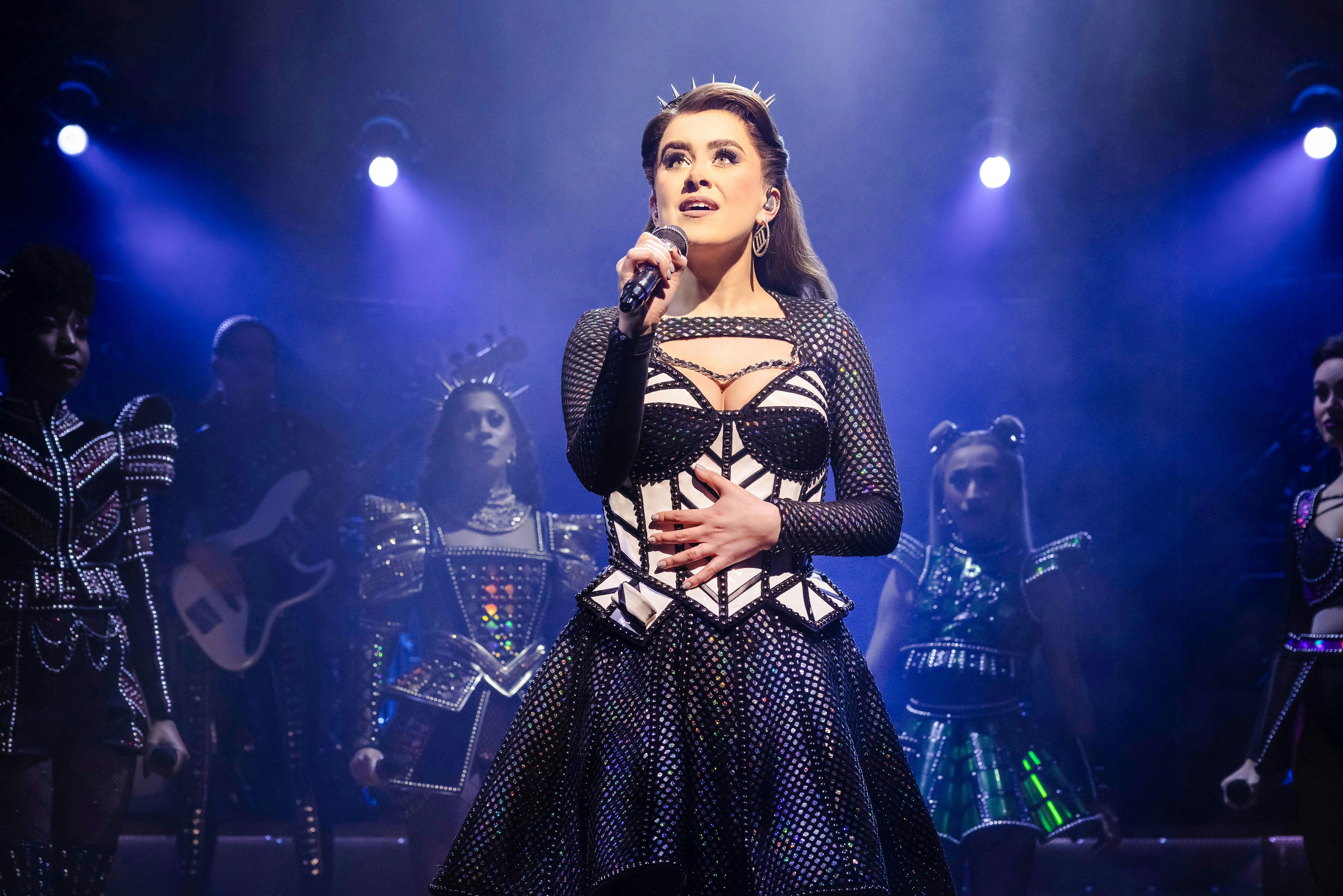 Performer singing on stage during the musical "Six" with a spotlight and backup singers.