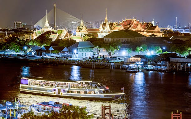 Cruise ship on Chao Phraya River with Grand Palace illuminated in the background at night, Bangkok.