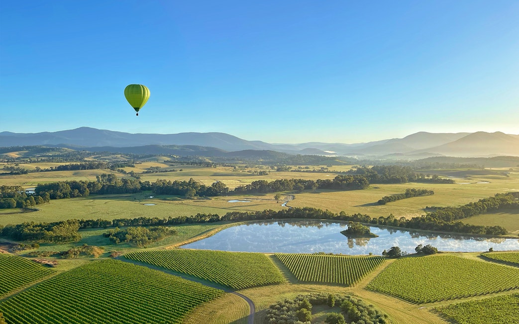 Hot air balloon over Yarra Valley vineyards at sunrise, with mountains in the background.