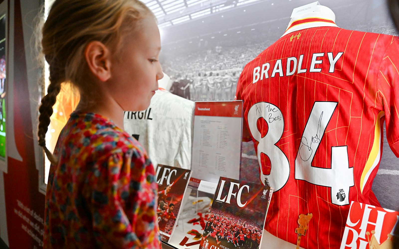 Young girl viewing signed Liverpool FC jersey at Anfield museum display.