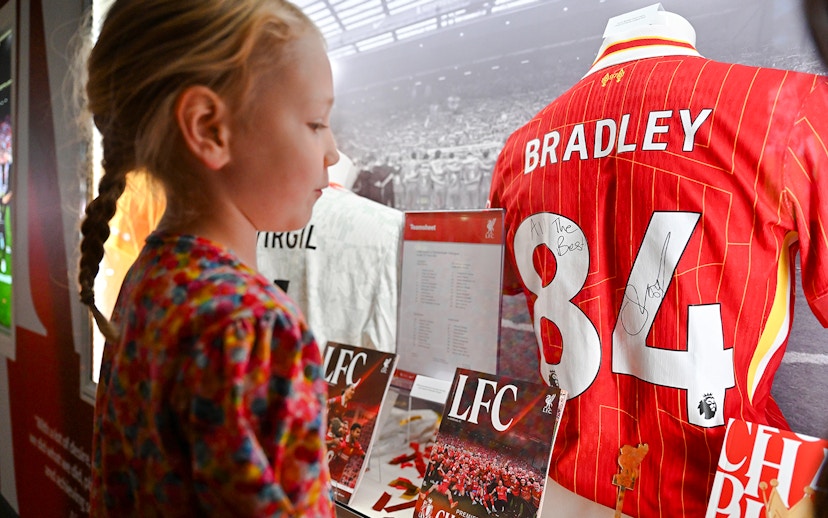 Young girl viewing signed Liverpool FC jersey at Anfield museum display.