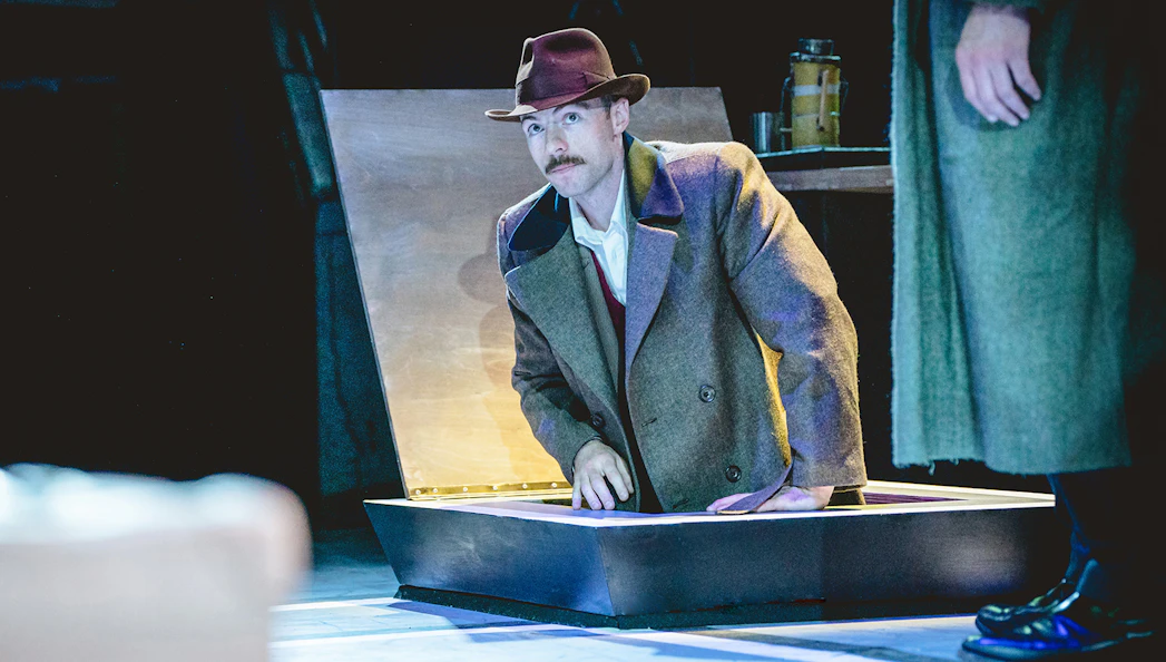 Man in a trench coat and hat emerging from a trapdoor on stage during Tom, Dick and Harry performance.