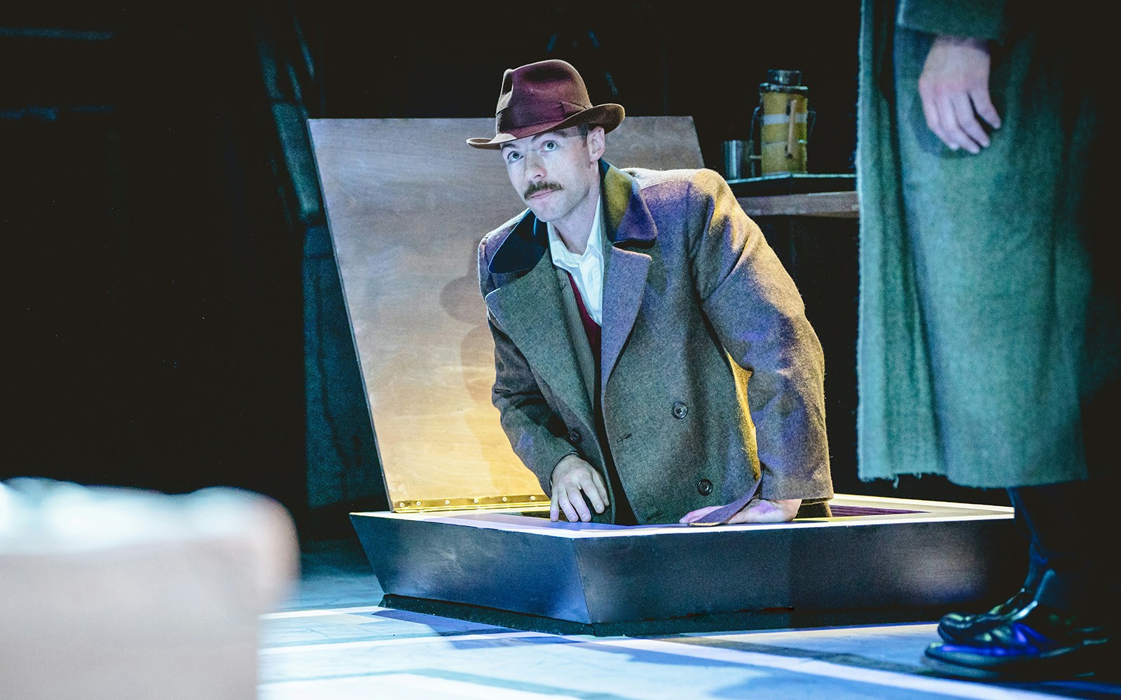 Man in a trench coat and hat emerging from a trapdoor on stage during Tom, Dick and Harry performance.