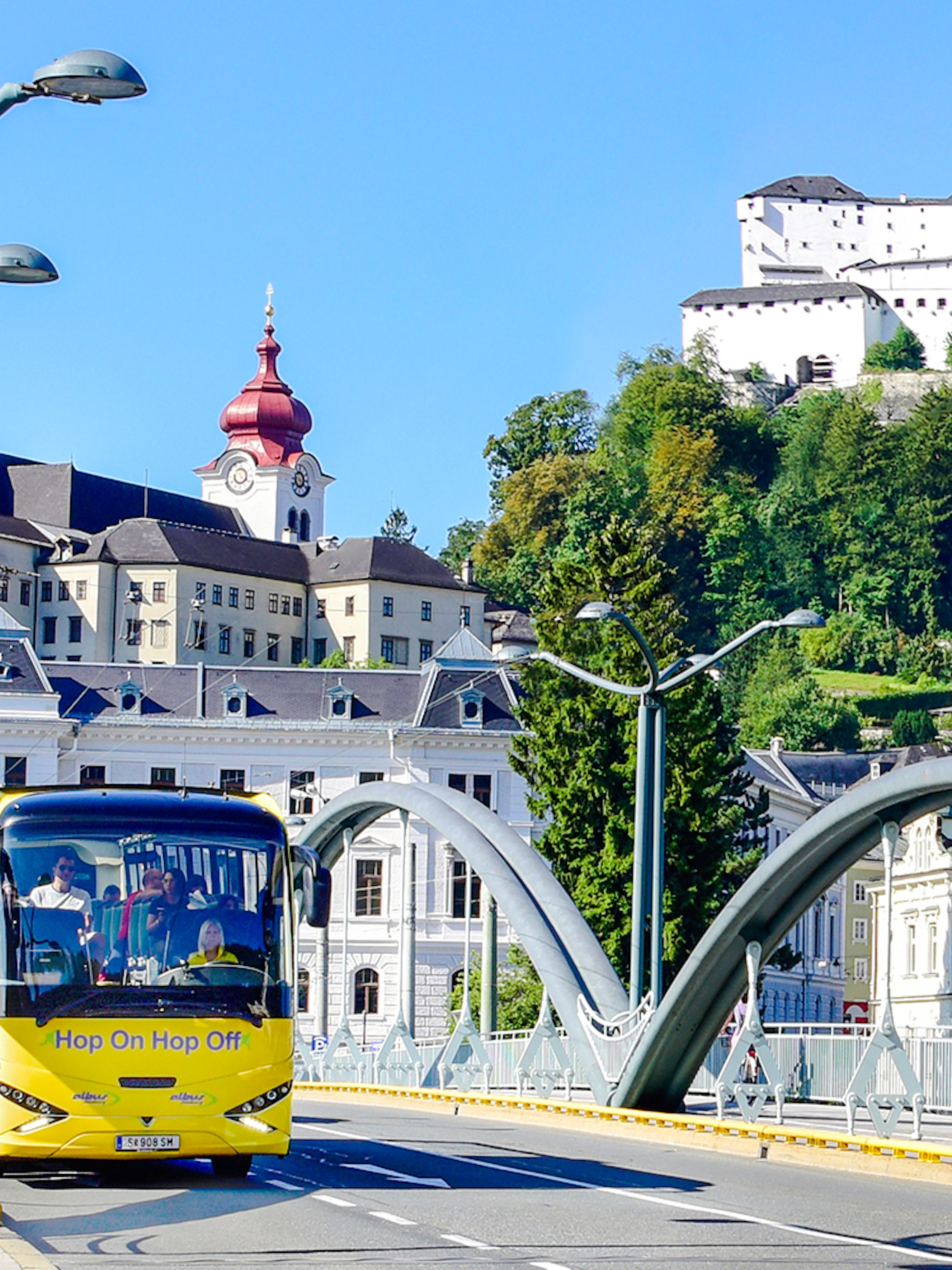 Hop On Hop Off bus crossing a bridge with Hohensalzburg Fortress in Salzburg.