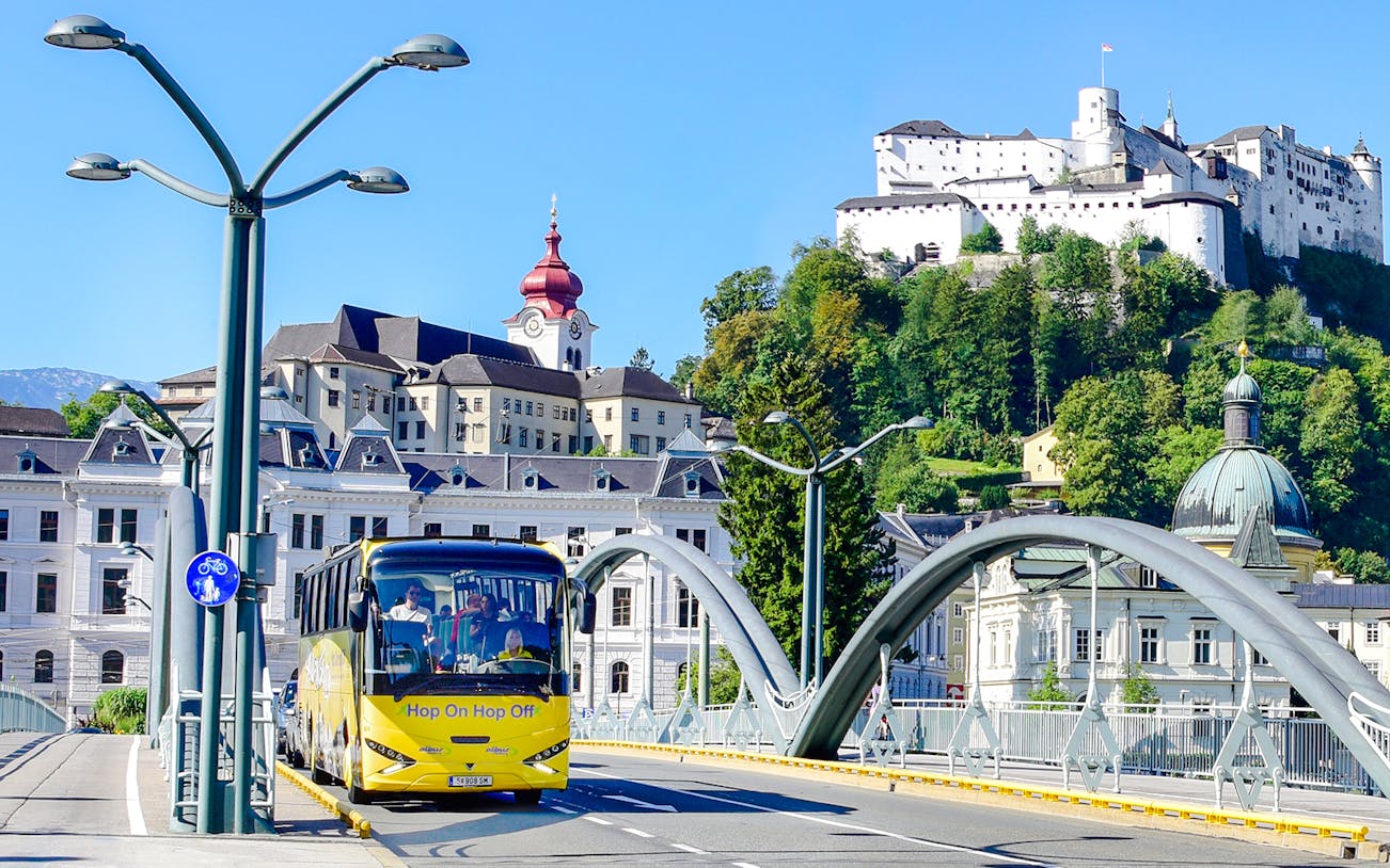 Hop On Hop Off bus crossing a bridge with Hohensalzburg Fortress in Salzburg.