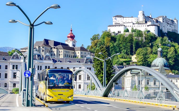 Hop On Hop Off bus crossing a bridge with Hohensalzburg Fortress in Salzburg.