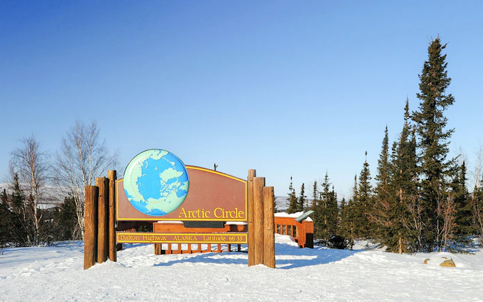 Arctic Circle sign on Dalton Highway, Alaska, surrounded by snow and trees.