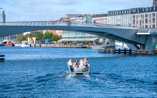 Guests on a boat tour in Copenhagen harbor, passing under a modern bridge.