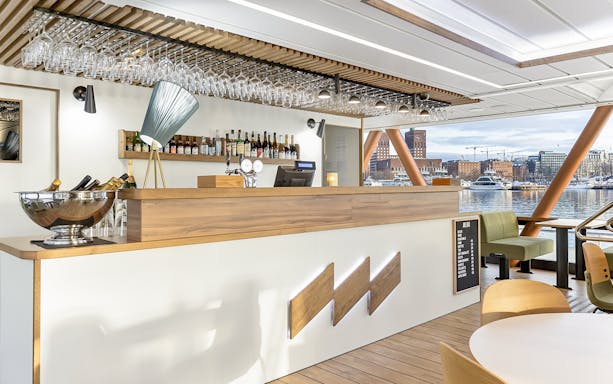 Bar area on a boat with Oslo waterfront view, featuring wine bottles and glasses.