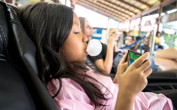 Child playing a game on a phone while blowing a bubble in an Asian travel setting.
