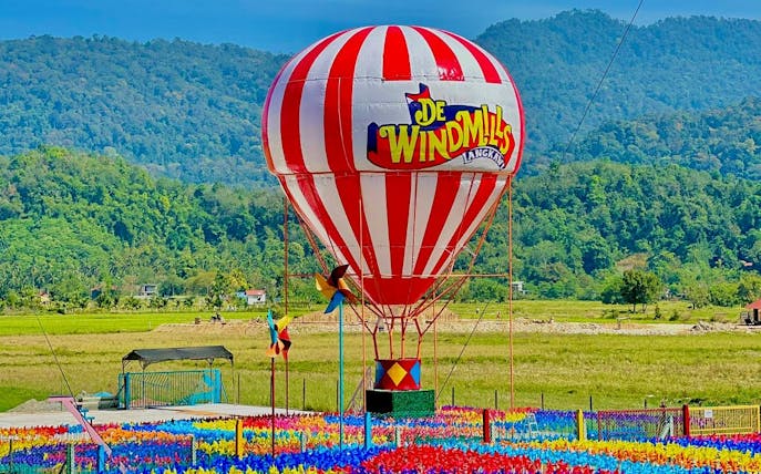 Hot air balloon at De Windmill Selfie Park, Langkawi, with colorful pinwheels and scenic mountain backdrop.