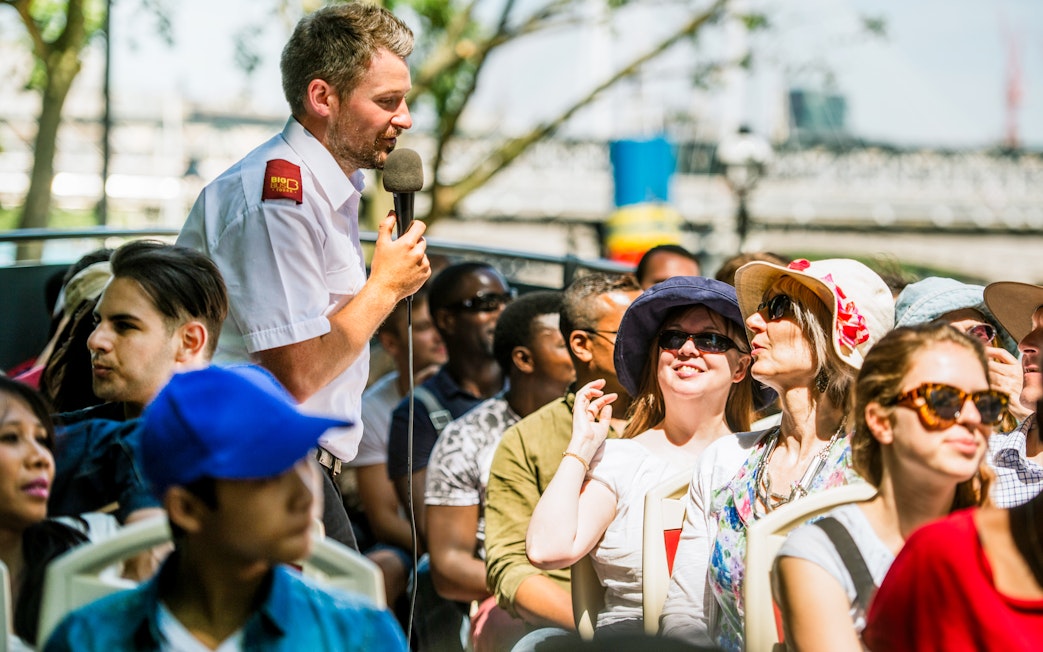 Tour guide speaking to passengers on an open-top bus tour in a city setting.
