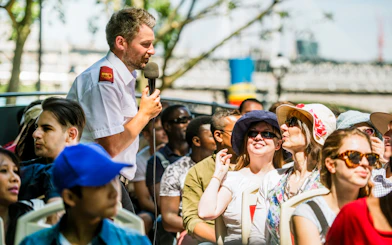 Tour guide speaking to passengers on an open-top bus tour in a city setting.